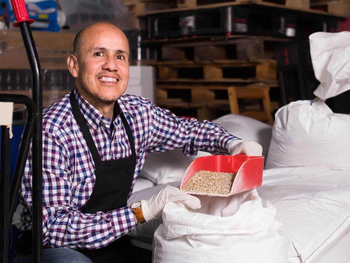 Man in plaid shirt and apron scooping brewers malt out of a white bag in a warehouse setting