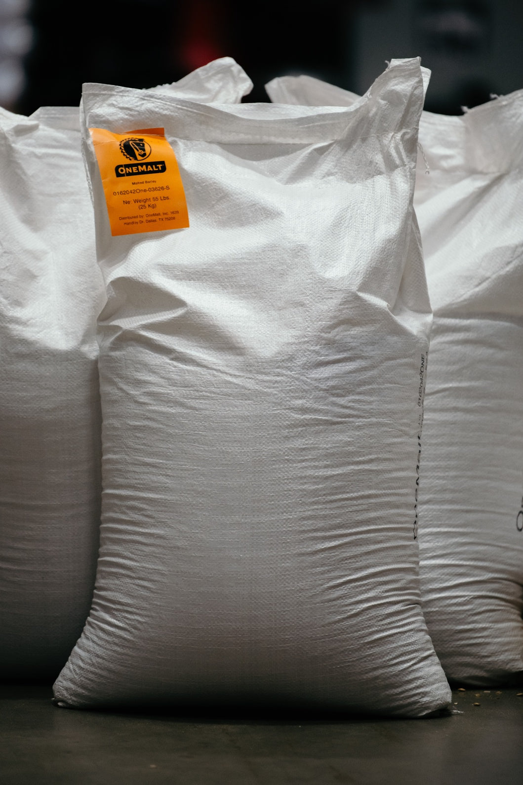 Three white bags of OneMalt malt product in a warehouse setting