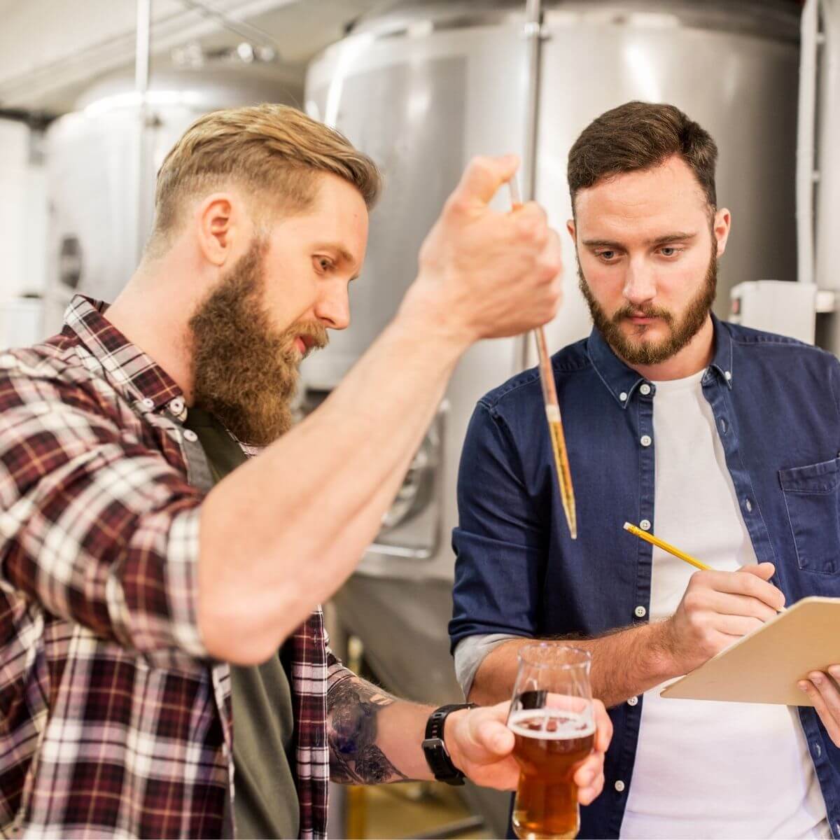 Two men in a brewery setting, one holding glass of beer and the other taking notes